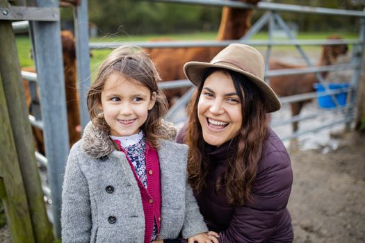 Brunette woman smiling at the camera while her smiling little daughter next to her, is looking away, and with brown alpacas behind them