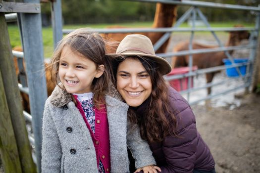 Brunette woman and her little daughter smiling and looking at the distance, while brown alpacas eat behind them