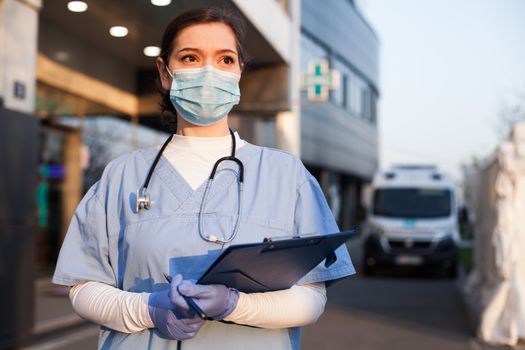 Young female EMS key worker doctor in front of healthcare ICU facility,wearing protective PPE face mask equipment,holding medical lab patient health check form,UK&amp;US COVID-19 pandemic outbreak crisis
