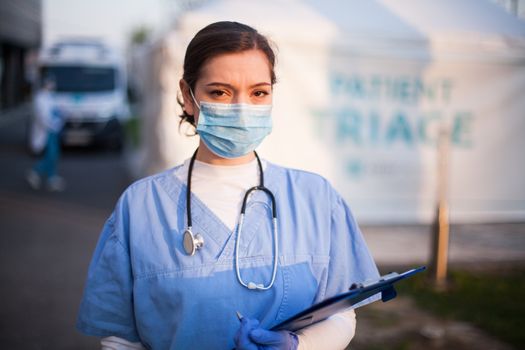 Portrait of very tired & exhausted UK NHS ICU doctor in front of hospital,emergency patient triage tent in background,Coronavirus COVID-19 pandemic outbreak crisis,medical staff working long shifts