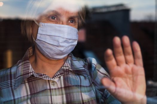 Elderly caucasian woman wearing hand made protective face mask, in nursing care home, looking outside window with sadness in her eyes, self isolation due to the global COVID-19 Coronavirus pandemic