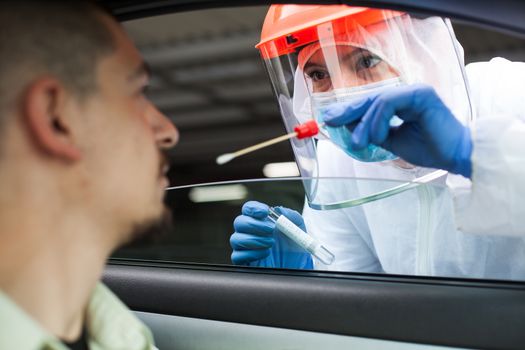 Medical UK NHS worker performing drive-thru COVID-19 test,taking nasal swab specimen sample from male patient through car window,PCR diagnostic for Coronavirus presence,doctor in PPE holding test kit