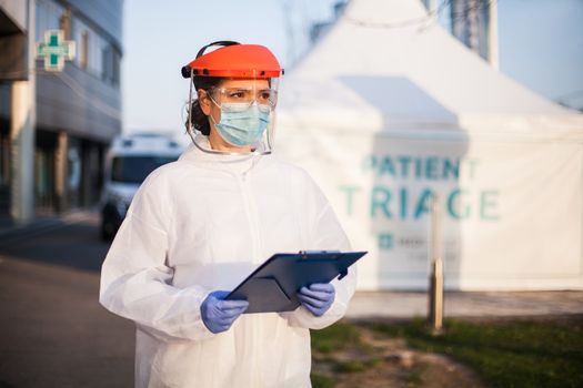 Paramedic wearing personal protective equipment PPE holding folder standing in front of ICU hospital isolation rt-PCR drive thru testing site,COVID-19 pandemic outbreak crisis,worried exhausted staff