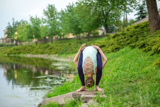 A young sports girl practices yoga on a green lawn by the river, assans posture. Unity with nature