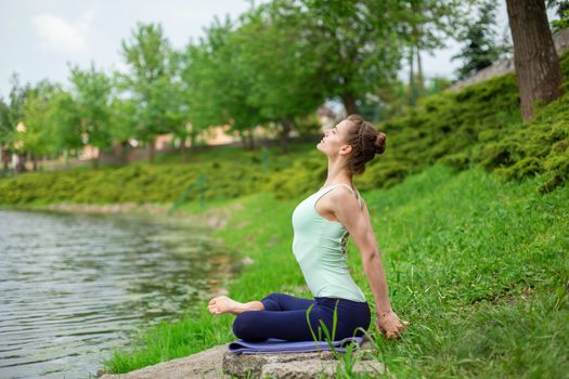 A young sports girl practices yoga on a green lawn by the river, assans posture. Unity with nature