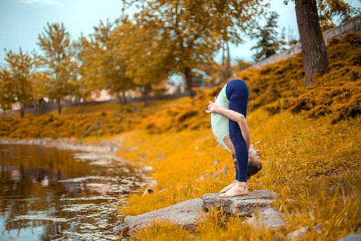 Slender young brunette yogi performs challenging yoga exercises on the green grass in autumn against the background of nature.