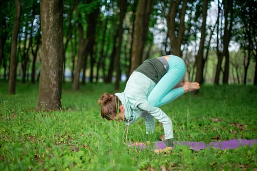 Thin brunette girl doing Crane exercise, Bakasana pose in a summer park. Green lush forest on the background. Woman doing exercises on a yoga mat.