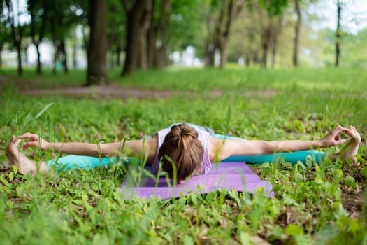 Thin brunette girl plays sports and performs beautiful and sophisticated yoga poses in a summer park. Green lush forest on the background. Woman doing exercises on a yoga mat.