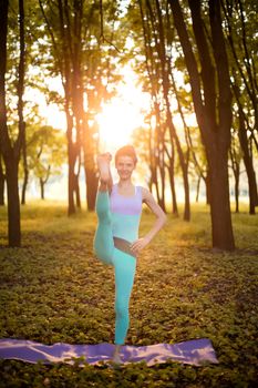 Thin brunette girl plays sports and performs yoga poses in autumn park on a sunset background. Woman doing exercises on the yoga mat. Autumn forest. Soft focus.