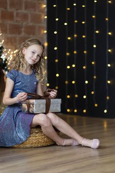 The adorable little girl in gorgeous dress holds gift box sitting near Christmas tree.