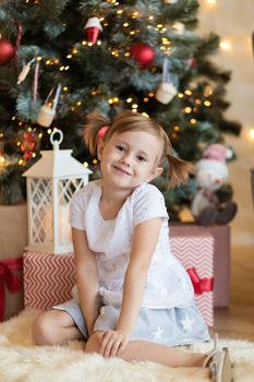Pretty little child is sitting in front of christmas tree among garlands and presents..
