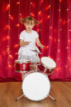 Little smiling girl plays on drums on red background with new year garlands.