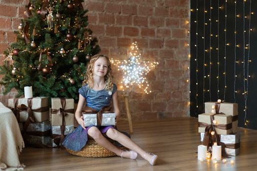 Pretty little child is sitting in front of christmas tree among garlands and presents..