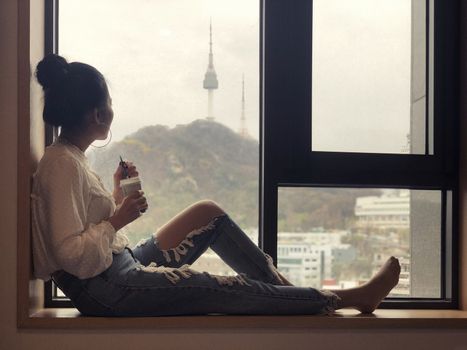 Young woman drinking coffee and looking out the window to see the view of Seoul Tower in the morning