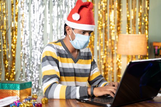 Young man in medical mask busy working on laptop during Christmas or new year celebration eve with decorated background and gifts in front - concept of work from home during holydays
