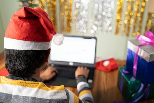 Shoulder shot of young man busy working on laptop with Santa hat during Christmas or new year eve - Concept of work from home during holiday season due to coronavirus or covid-19 pandemic.