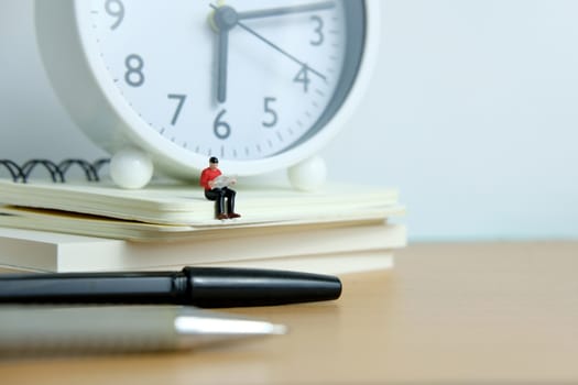 Young man sitting on book pile while reading with pen and white clock