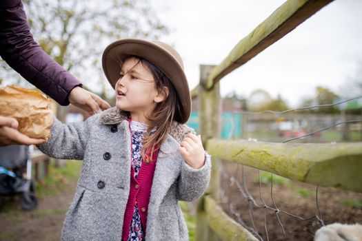 Happy little girl wearing cowboy hat interacting with her mom on a farm. Adventurous girl preparing to feed farmyard animals. Feeding barnyard animals