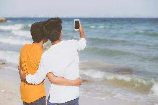 Back view gay portrait young couple smiling taking a selfie photo together with smart mobile phone at beach, LGBT homosexual lover in the vacation at sea, two man going to travel, holiday concept.