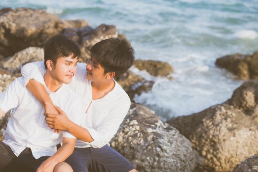 Homosexual portrait young asian couple sitting hug together on rock or stone in the beach in summer, asia gay going tourism for leisure and relax with happy in vacation at sea, LGBT legal concept.