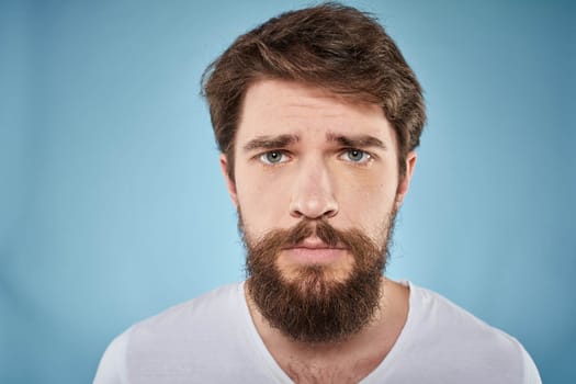 Bearded man displeased facial expression emotions close-up blue background white t-shirt. High quality photo