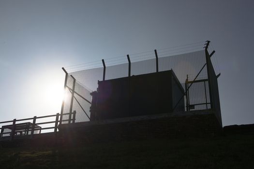 Tall barbed wire anti climb fence encases large square metal industrial water management equipment. Looking up, plain blue sky and bright sun behind