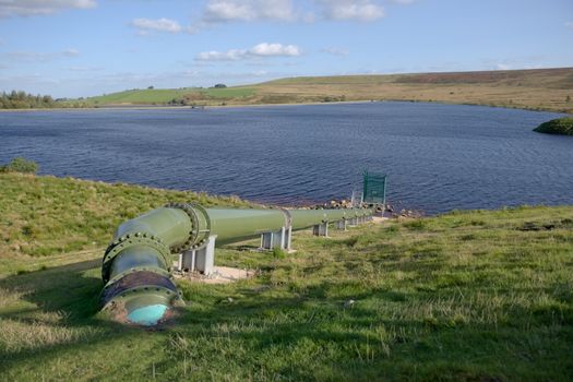 Warm sunlight shines on a shiny green pipe feeding water into a deep blue rural reservoir. Cloudy sky background. Modern industrial hydro equipment.
