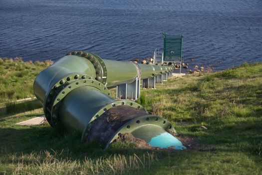 Full frame image of large green metal pipe in an outdoor hydro dam setting. Modern and bolted together, with control equipment at the reservoir end