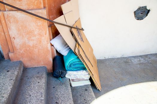 The possessions of a homeless person at his sleeping place with blankets, mattresses, cardboard and pillows on a wall in front of a staircase.