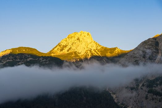 A mountain with mountains, which is illuminated by the sun and where in the shady foreground is still fog