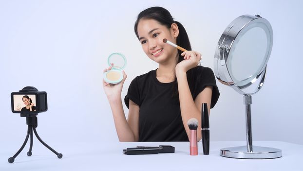 Asian teen woman sit in front of camera and live broadcasting as a beauty blogger influencer or youtuber to review or advice about how to make up at home. studio shot white background.