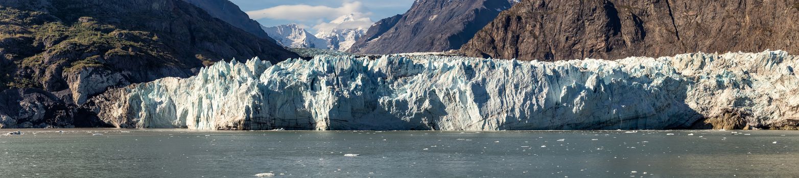 Panoramic view of Margerie glacier in Glacier Bay National Park and Preserve, Alaska, United States. Snowy mountain peaks and cloudy blue sky in the background.