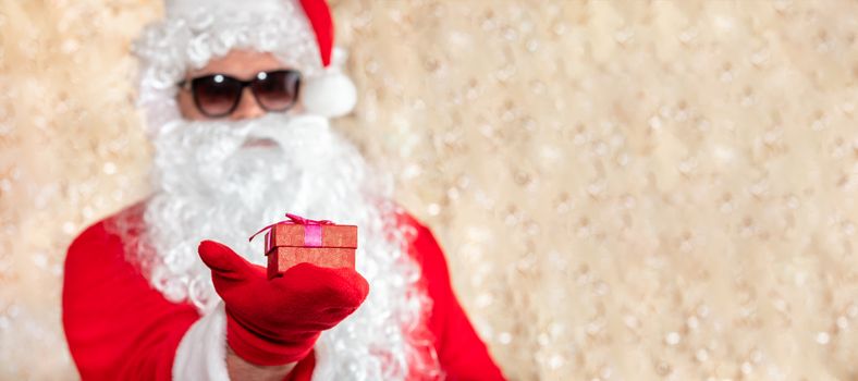 Santa Claus holding a Christmas present, red box with ribbon. Santa wearing sunglasses, with a long white beard, and is out of focus. Golden blurred sparkling background with copy space. Banner size.
