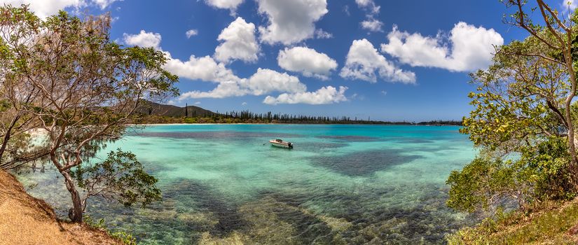 Amazing panoramic shot of Isle of Pines in New Caledonia. Beautiful turquoise water of the ocean with trees on the banks. Lone fishing boat anchored in the middle. Blue cloudy sky as a background.