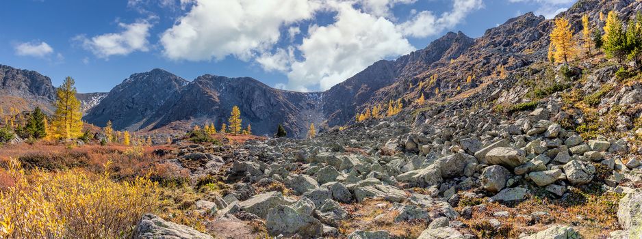 Altai mountains. Beautiful highland autumn panoramic landscape. Rocky foreground with golden trees. Blue cloudy sky as a background. Russia. Siberia.