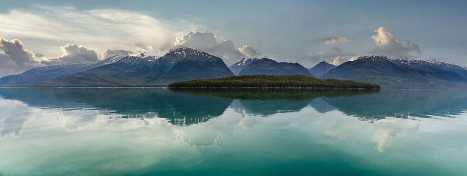 Beautiful landscape of snow capped mountains and a lone island full of green trees in a fjord in Alaska, USA. Cloudy sunset sky as a background. Mountains and sky reflecting in the water.