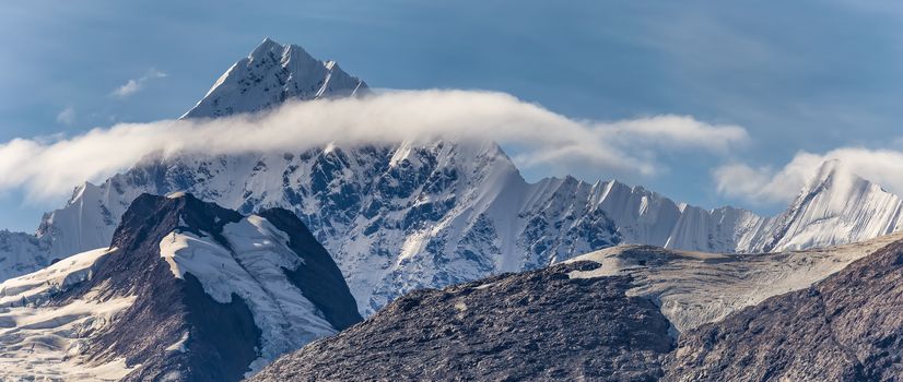 Beautiful panoramic view of a mountain peak covered with ice and snow, surrounded with fluffy couds in Alaska. Blue sky as a background.