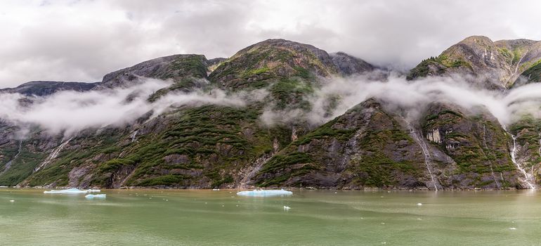 Beautiful panoramic view of mountains covered with fluffy clouds and trees in Alaskan fjord. Cloudy sky as a background.