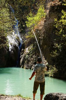 man catches fish in a mountain lake with a waterfall, rear view