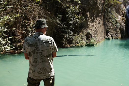 man catches fish in a mountain lake with a waterfall, rear view