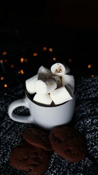 Mug with marshmallows and chocolate chip cookies on dark Christmas background with lights - vertical photo