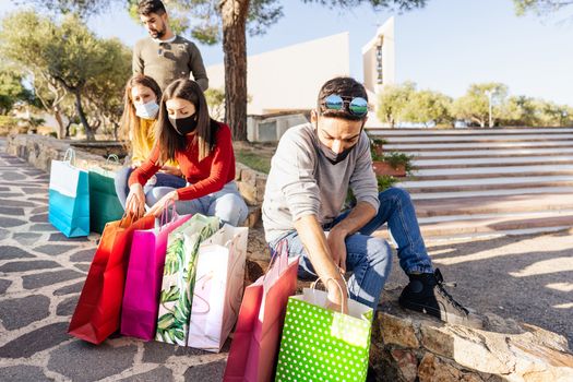 Caucasian young friends group sitting on a wall of the city square they rest after shopping wearing covid-19 coronavirus face masks looking into their colored shopping bags - Focus on man with glasses