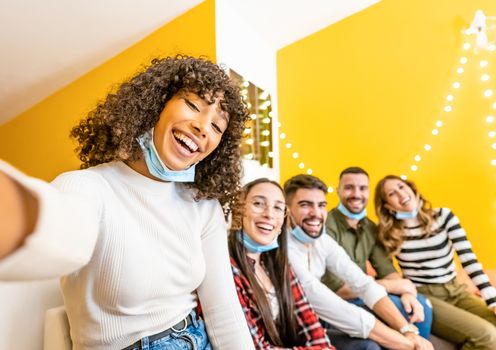 Group of multi ethnic young people having tech fun home - Beautiful black Hispanic woman take a photo with smartphone to her friends wearing lowered Coronavirus face mask protection - Focus on curly