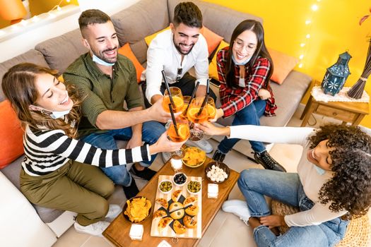 Group of young people toasting for home aperitif - Mixed race friends drinking spritz cocktail sitting on the sofa with snacks wearing lowered Covid-19 Coronavirus mask protection - Focus on glasses