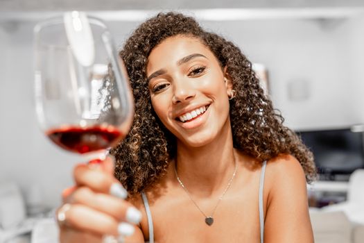 Happy young Hispanic curly brunette toasting with red wine glass looking at the camera in video call conference - Beautiful black woman sitting in living room chatting online with new social networks