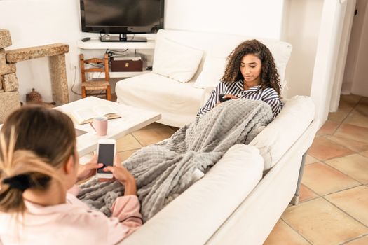 Mixed race female couple resting under a blanket on the sofa using smartphone to contacts friends online with social networks - Two young women technology addicted surfing the web - Focus on curly one
