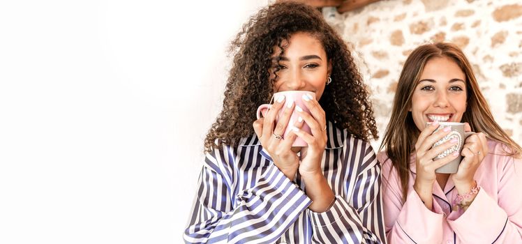 Mixed race female couple in pajama just woken up standing outdoor drinking tea looking at the camera - Two women friends smiling holding a mug with large white copy space to the left