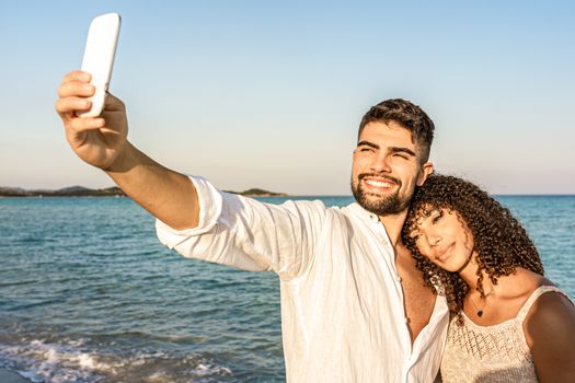 Mixed race couple in vacation at sea resort making a self portrait at sunset or dawn - Handsome smiling bearded man take photo with her Hispanic beautiful curly brunette girlfriend in loving position