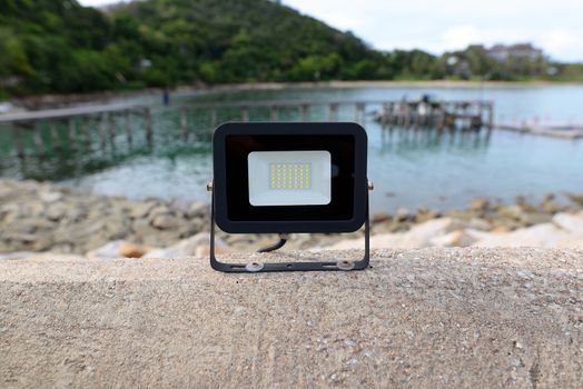 The black LED lamp is on the cement floor. With the sea shore as a blurred background.