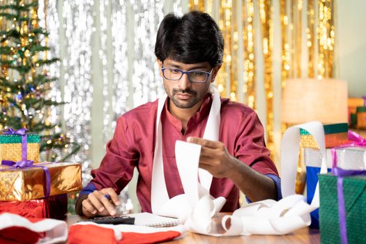 Young man busy in calculating holyday expenses after Christmas or new year 2021 holiday celebration showing with decorated background with gift in front.